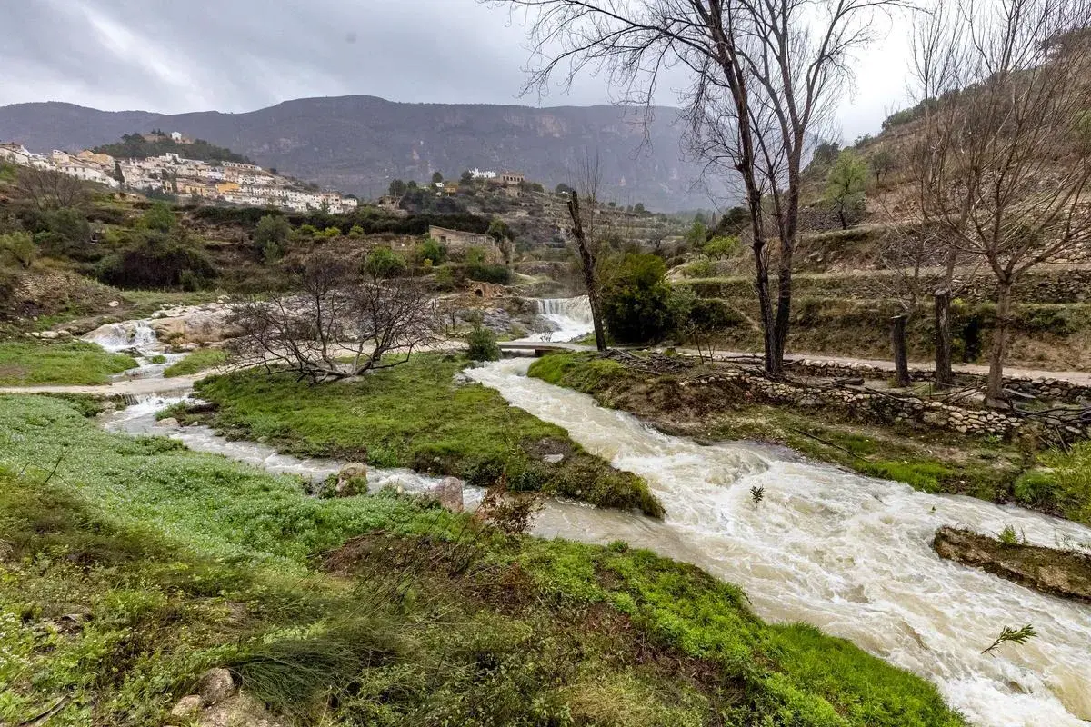 Río caudaloso cerca de Sella, Alicante, con cascadas y un pueblo blanco en la ladera.
