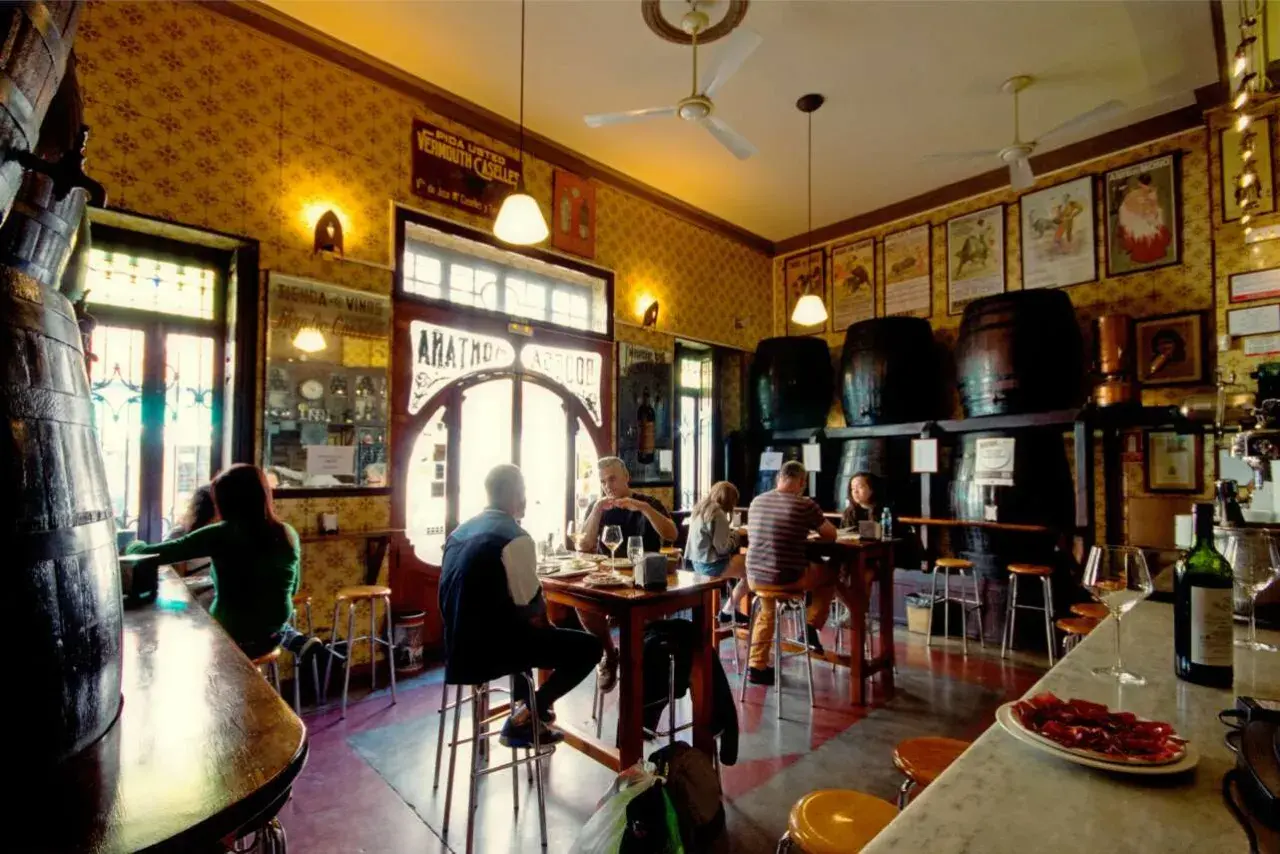 Gente en un bar tradicional, con barriles y carteles antiguos. Un lugar con encanto, como una casa de montaña en Valencia, donde disfrutar de tapas y vermut.