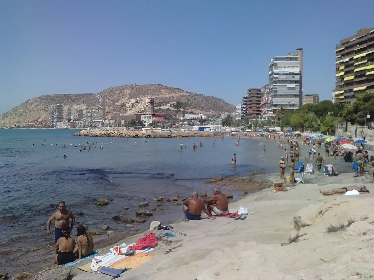 Gente disfrutando del sol y el mar en la playa de la Almadraba, con edificios modernos al fondo.