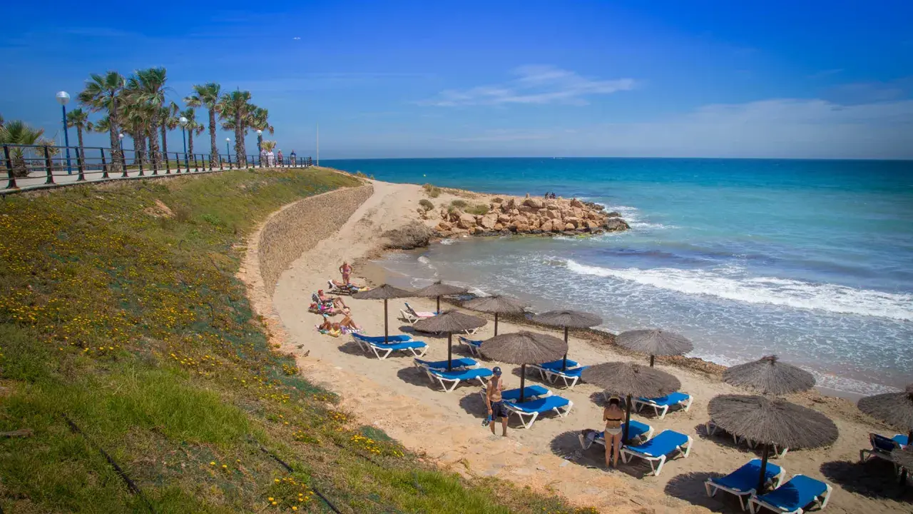 Una cala estaca con palmeras y sombrillas en la arena, donde la gente descansa bajo el sol. El mar azul turquesa invita a un refrescante baño.