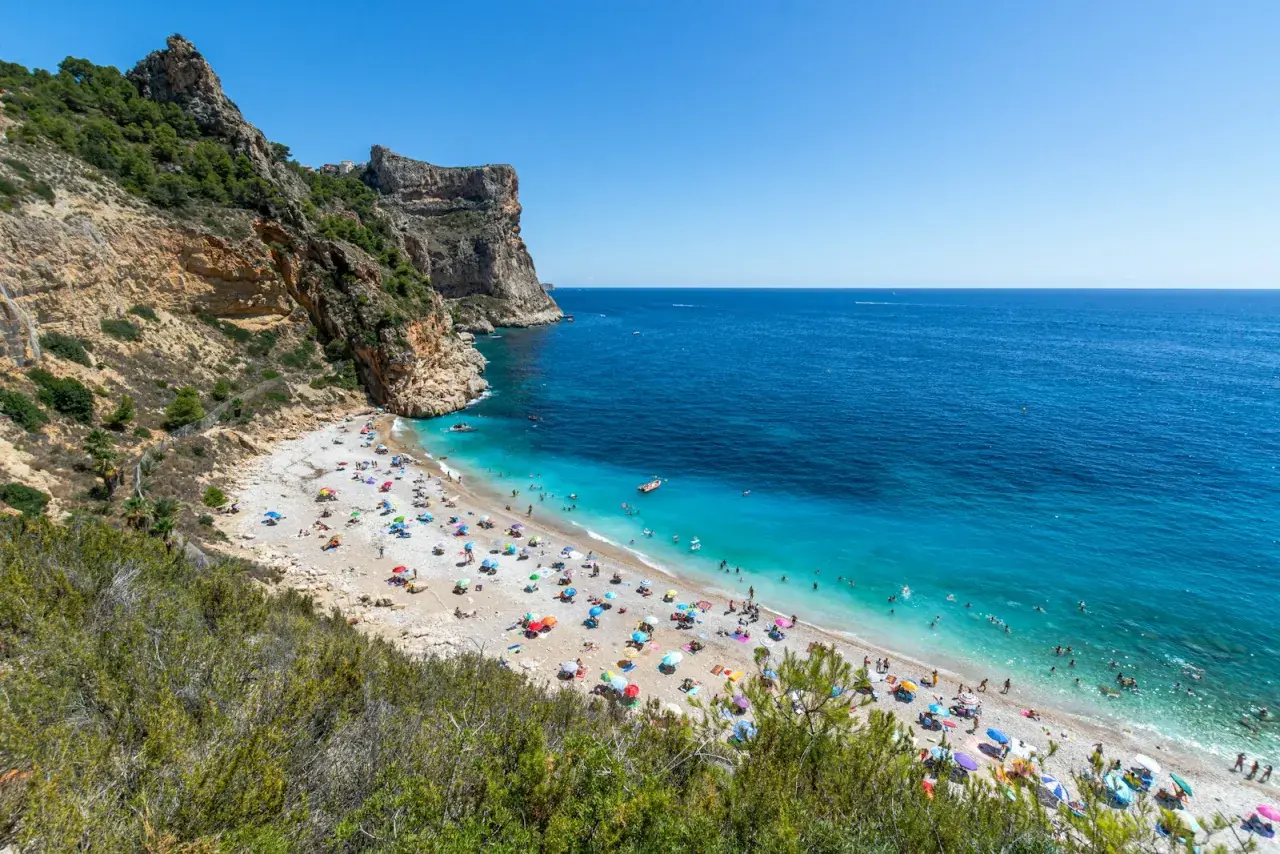 La **cala Moraig** es un paraíso de guijarros y aguas turquesas, con acantilados imponentes y gente disfrutando del sol bajo sombrillas de colores.