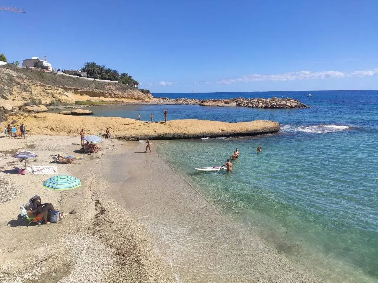 Gente disfrutando del sol y el mar en la cala Almadraba, Campello. Aguas cristalinas y rocas emergiendo del azul.