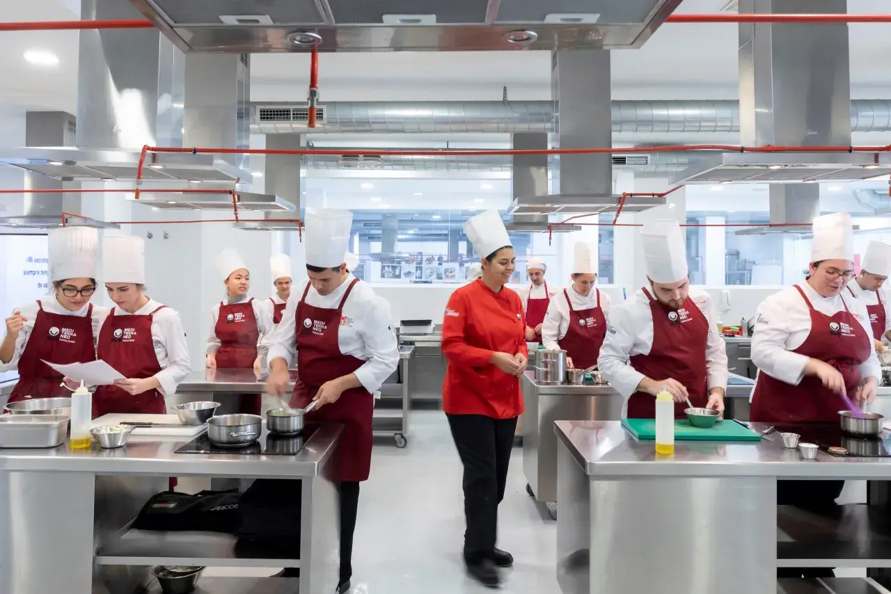 Estudiantes en un centro culinario mediterráneo, con toques rojos y blancos, aprendiendo a cocinar.