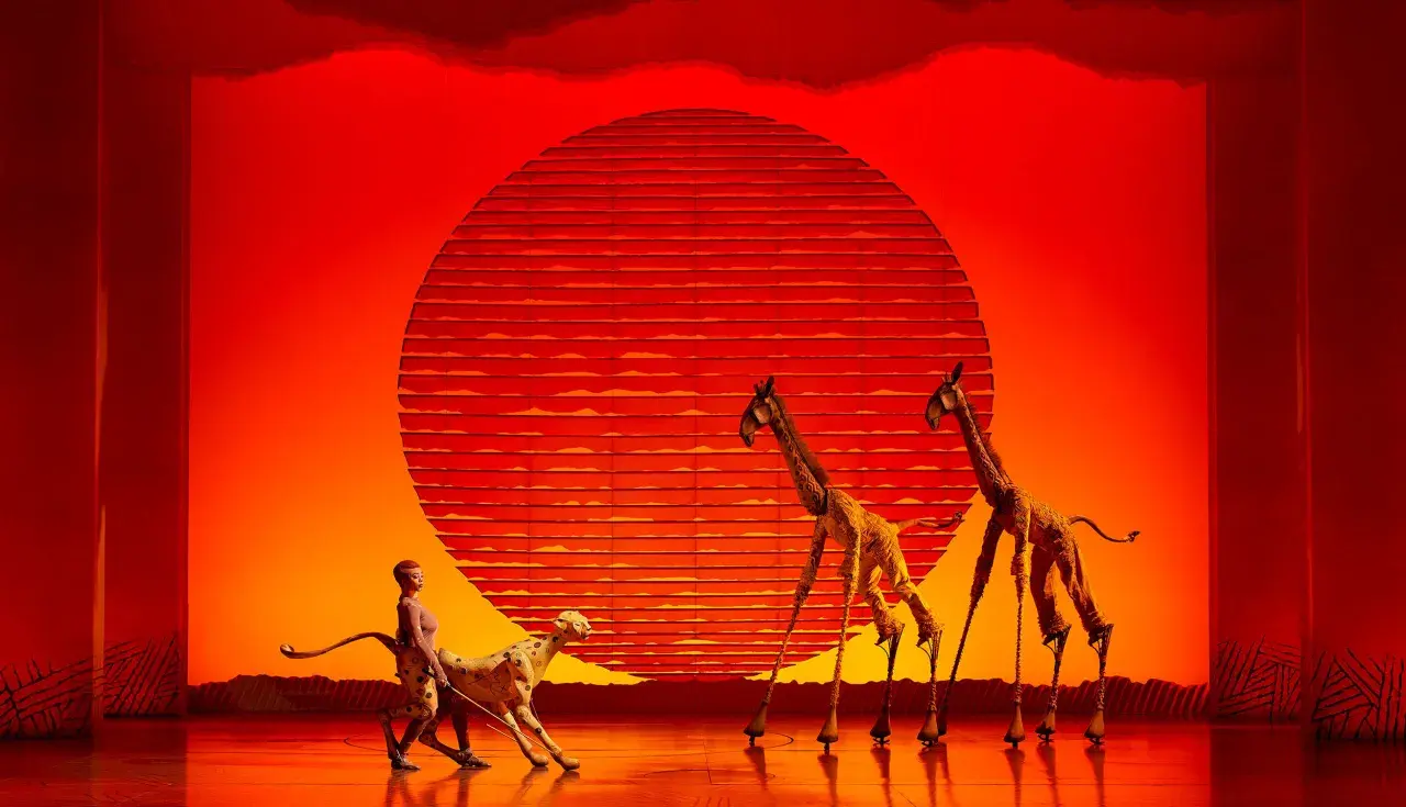 Escena de teatro en Valencia con un guepardo y jirafas con zancos ante un sol naranja.