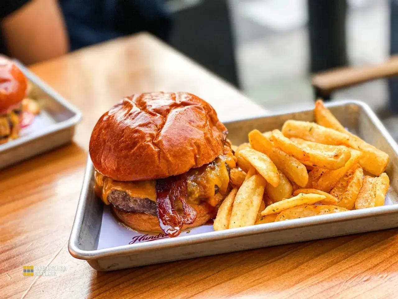 Jugosa hamburguesa con queso fundido y tocino crujiente, acompañada de patatas fritas doradas. Un festín para los sentidos en Hundred Valencia.