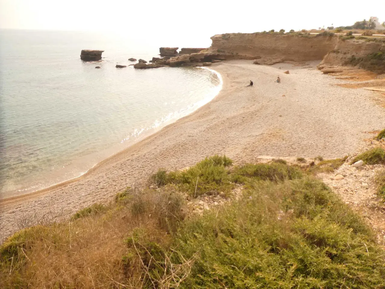 Una tranquila cala foradada con aguas cristalinas y rocas emergiendo del mar. Dos personas disfrutan de la soledad en la playa de guijarros.
