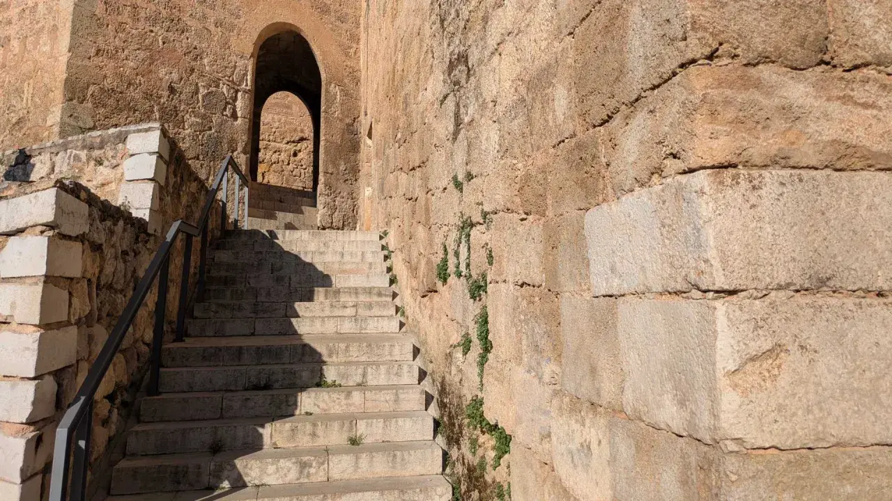 Escaleras de piedra ascienden hacia un arco en el castillo de Requena. La antigua muralla de piedra, con vegetación creciendo en sus grietas, proyecta sombras.