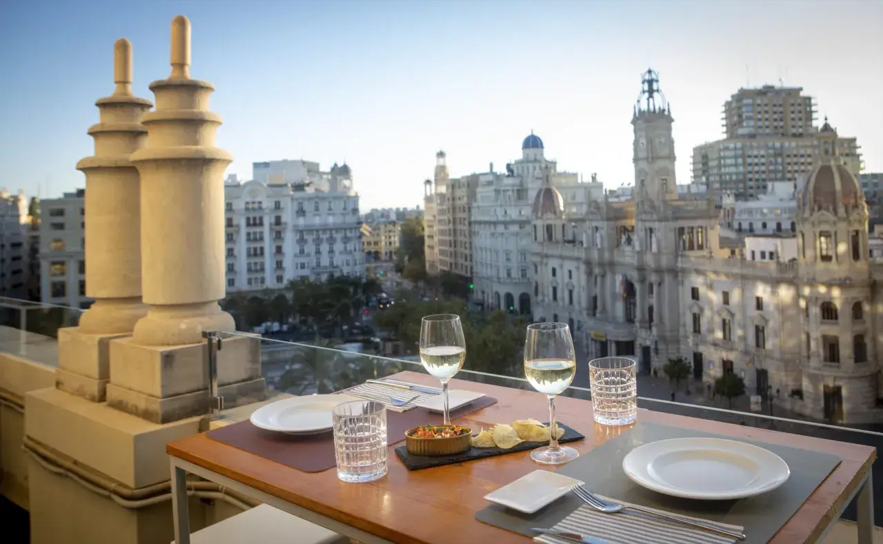 Una mesa en una terraza con vistas a la ciudad, con copas de vino y aperitivos, bajo el cielo de Atenea.