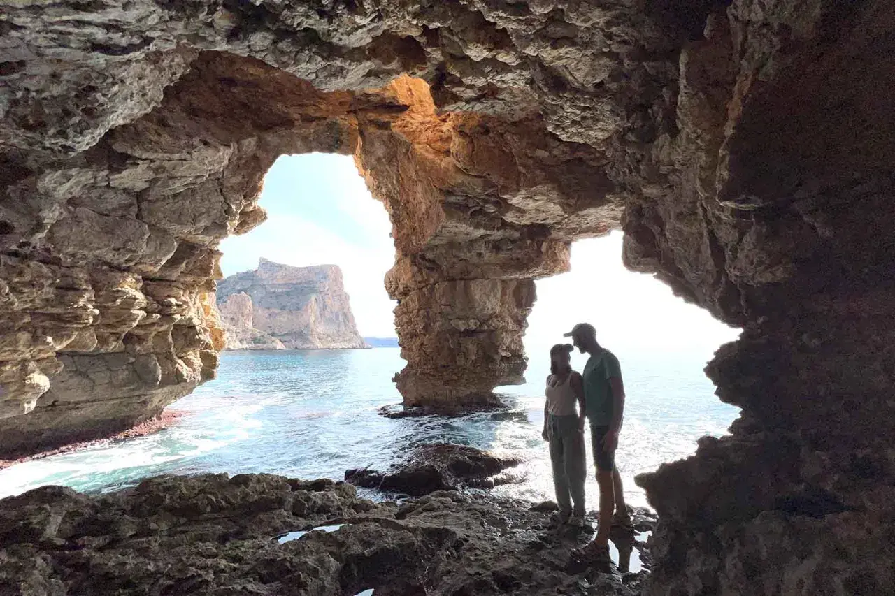 Pareja contemplando el mar desde la Cova dels Arcs, con sus impresionantes formaciones rocosas y el sol filtrándose.