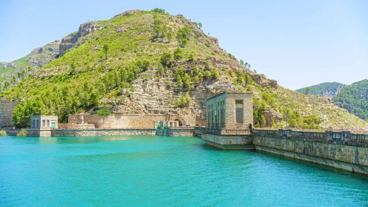 El embalse de Benagéber, con sus aguas turquesas y la imponente presa de piedra, se funde con la montaña verde.