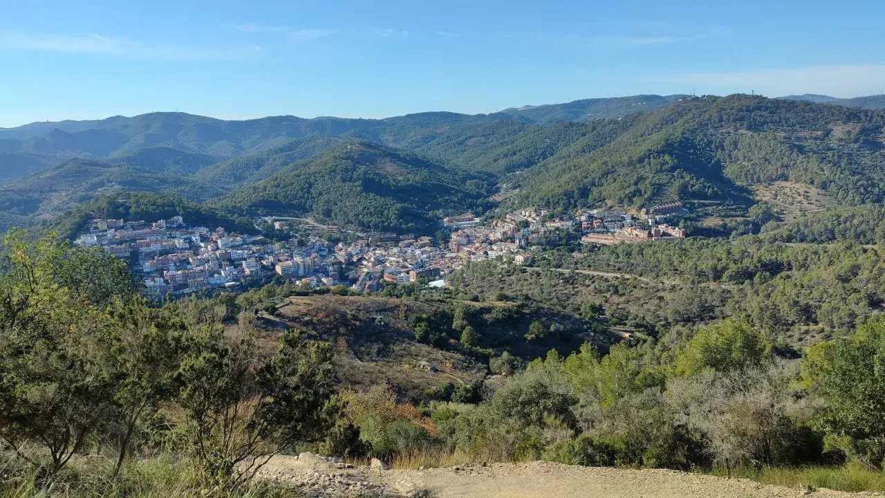 Vista panorámica de un pueblo en un valle rodeado de montañas boscosas. Se distingue la ermita de Sant Ramon.