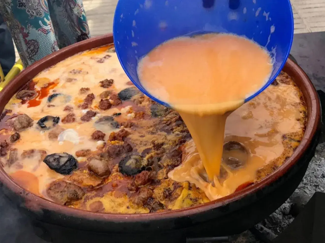 Un líquido naranja se vierte de un recipiente azul a una paella con carne y marisco.