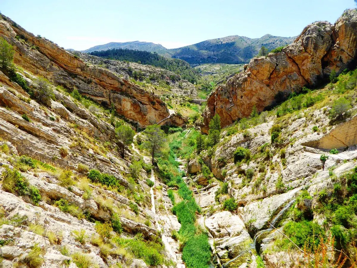 Un cañón rocoso con vegetación exuberante y un río serpenteante. El **pantano de Tibi** se revela en su esplendor natural, con formaciones geológicas impresionantes y un cielo azul claro.