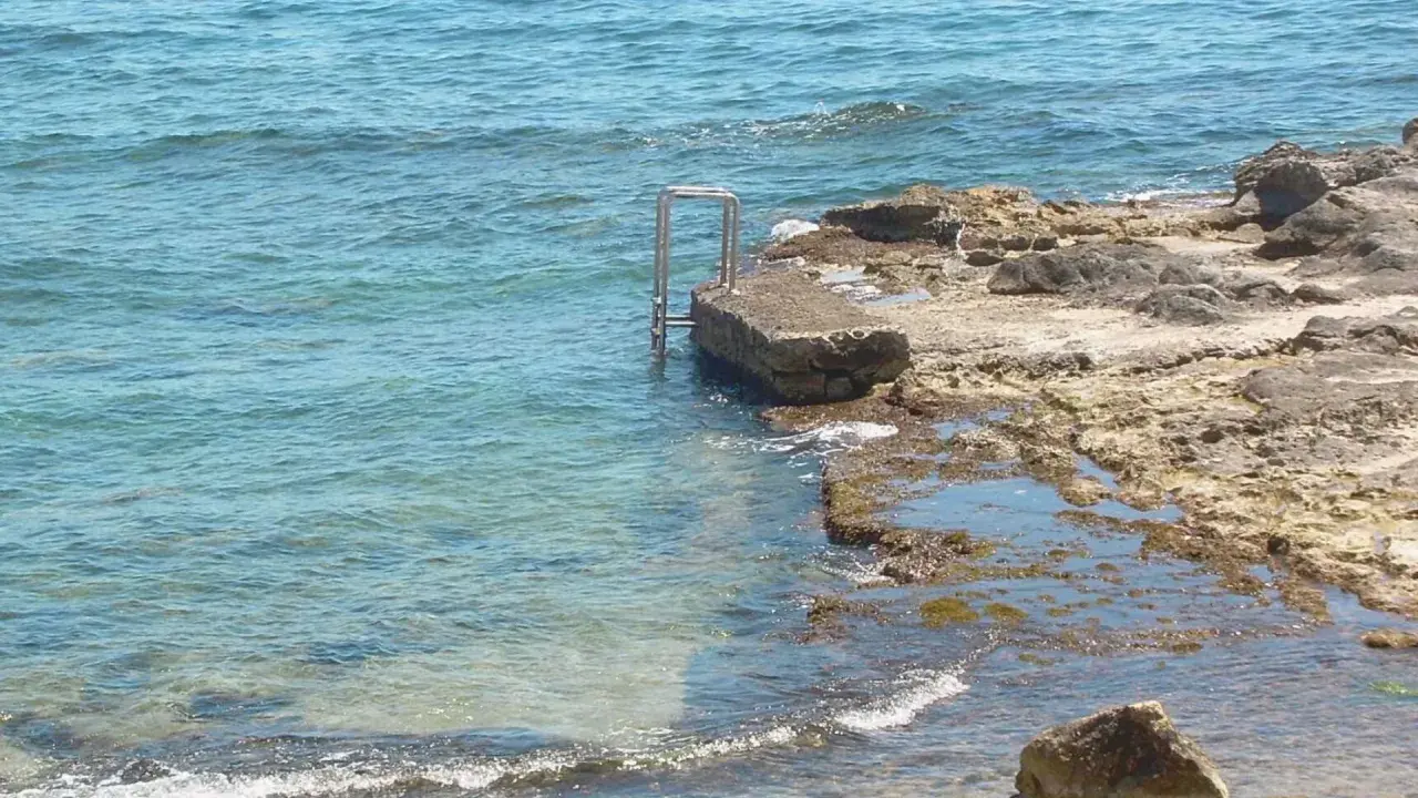 Escalera metálica en una roca que se adentra en el mar azul de cala l'andragó.
