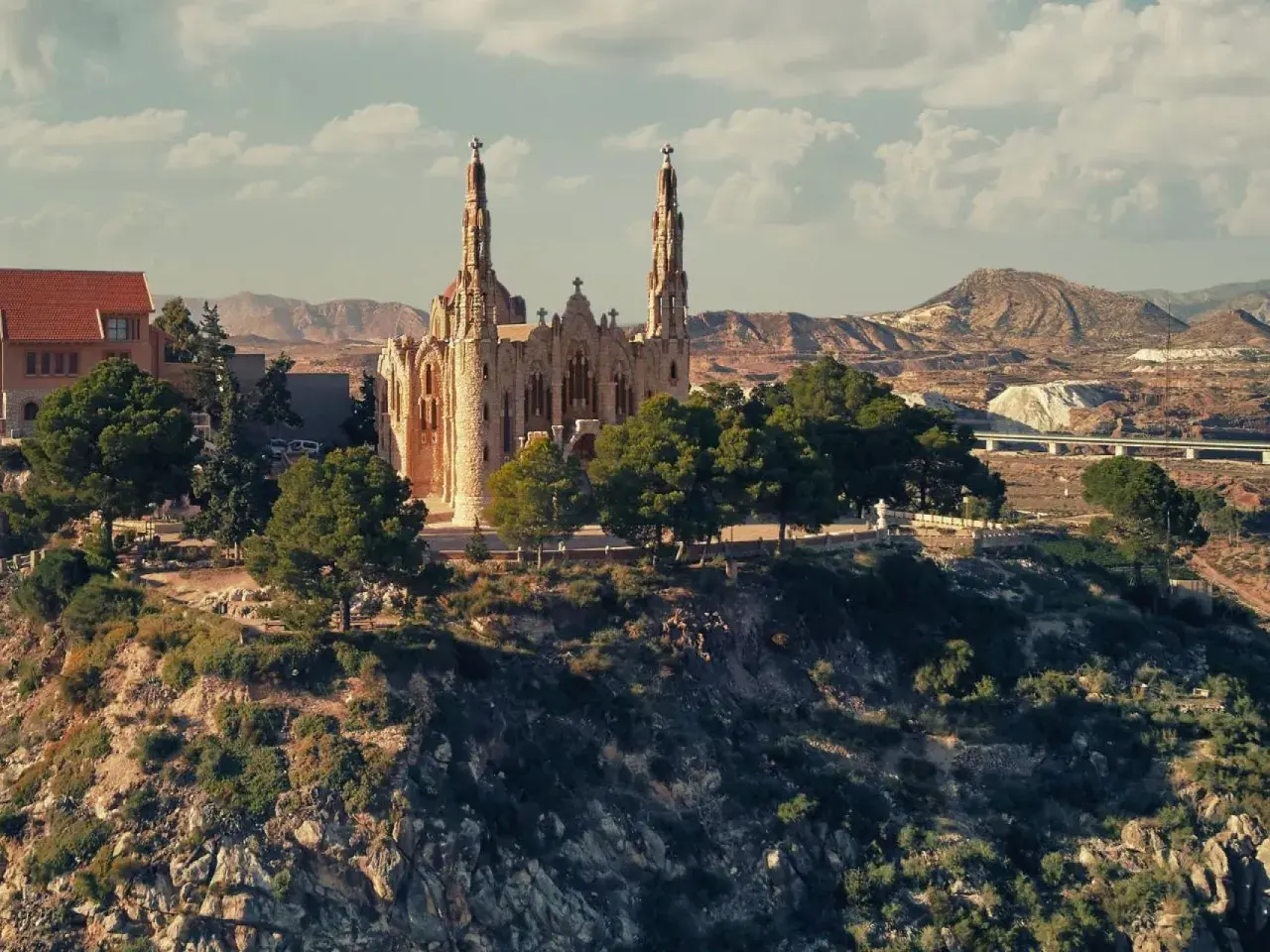 El **santuario** de Novelda se alza majestuoso sobre una colina, rodeado de vegetación y con un paisaje árido de fondo.