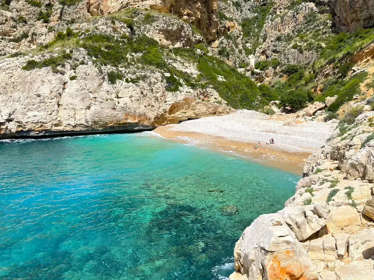 Aguas turquesas bañan la cala testos, una playa de guijarros rodeada de acantilados rocosos y vegetación.