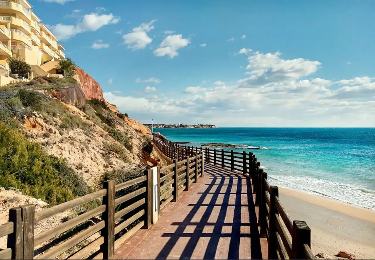 Paseo de madera con vistas a las mejores playas de Cabo Roig. Mar turquesa, arena dorada y un edificio amarillo en la ladera.