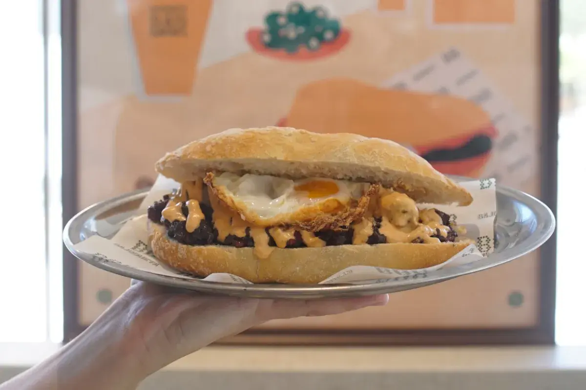 Un delicioso almuerzo valenciano: un bocadillo con carne, huevo frito y salsa cremosa, servido en plato metálico.