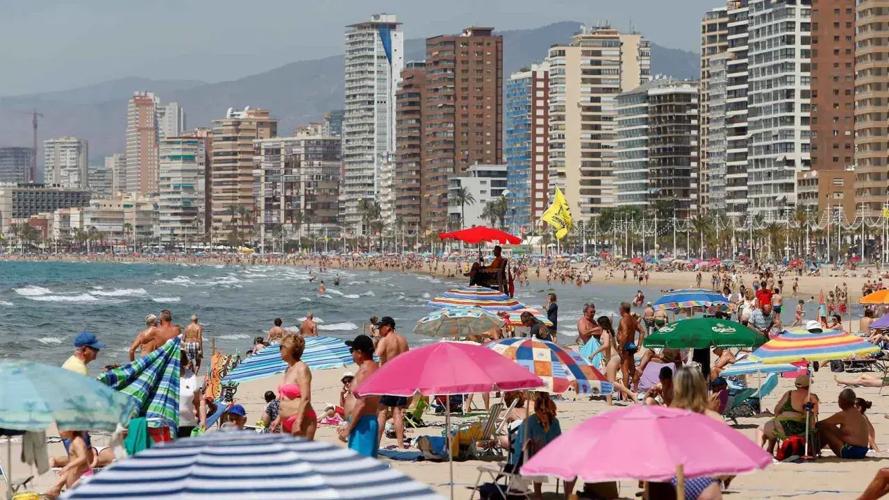 Gente disfrutando de un día soleado en la mejor playa de Benidorm, con edificios altos de fondo y sombrillas de colores.