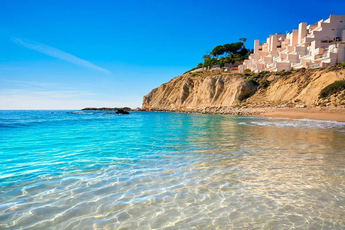 Aguas cristalinas en las calas de Alicante, con una urbanización blanca en la cima del acantilado y el mar azul intenso.