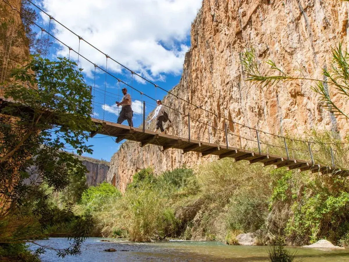Dos excursionistas cruzan un puente colgante sobre un río, explorando las rutas de senderismo de Valencia.