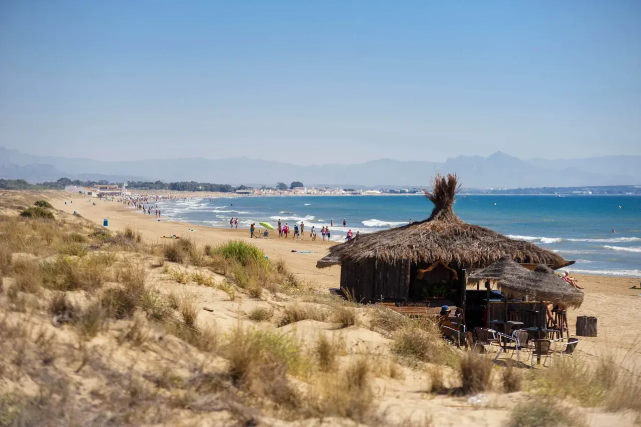 Chiringuito de paja en la playa del Rebollo, con gente disfrutando del sol y el mar azul. Montañas al fondo.