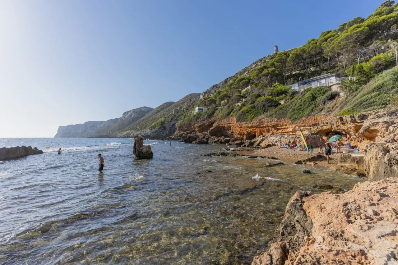 Gente disfruta del sol y el mar en una cala rocosa. Se ven casas y una torre en la **punta negra** de la colina verde.