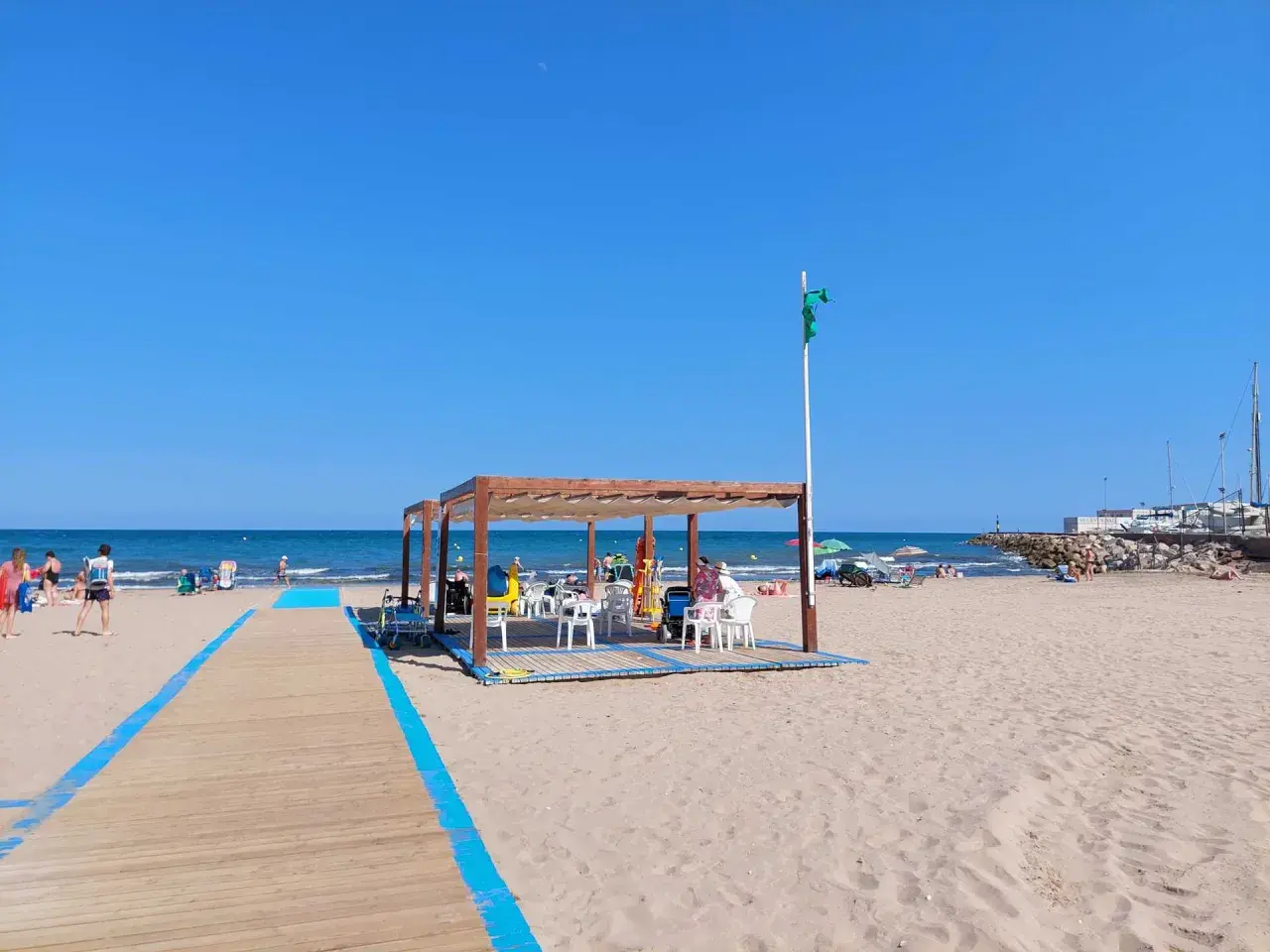 Pasarela de madera azul en la playa de Massamagrell, con chiringuito y gente disfrutando del sol y el mar.