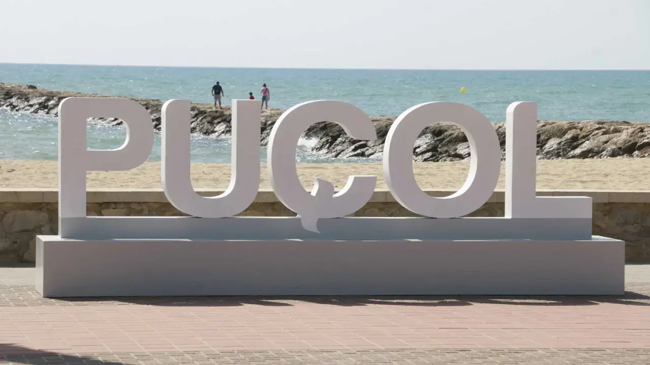 Letras gigantes "PUZOL" en la playa, con el mar azul y personas al fondo.
