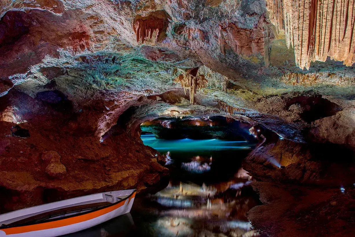 Interior de cueva con estalactitas, agua turquesa y una barca. Consulta las cuevas de Vall d'Uixó horarios y precios para tu visita.