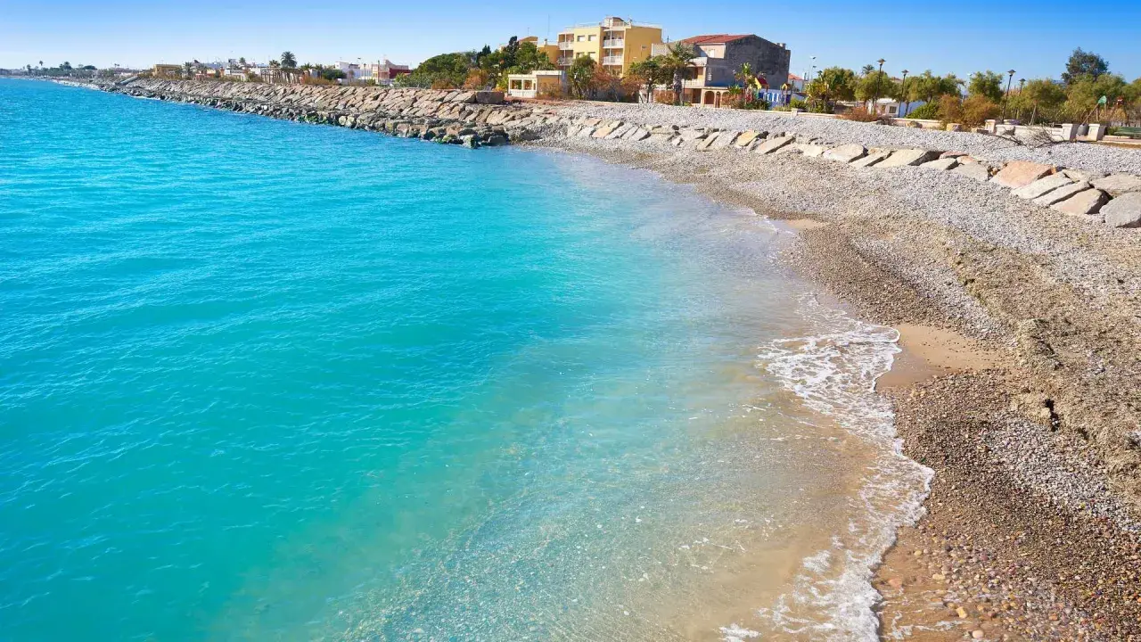 Aguas turquesas bañan la playa almenara, con un rompeolas de rocas y edificios al fondo.