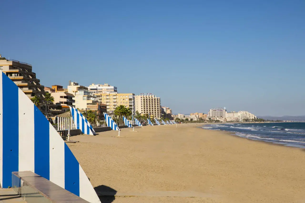 Amplia playa del Moro con edificios turísticos al fondo y sombrillas a rayas azules y blancas alineadas en la arena dorada.