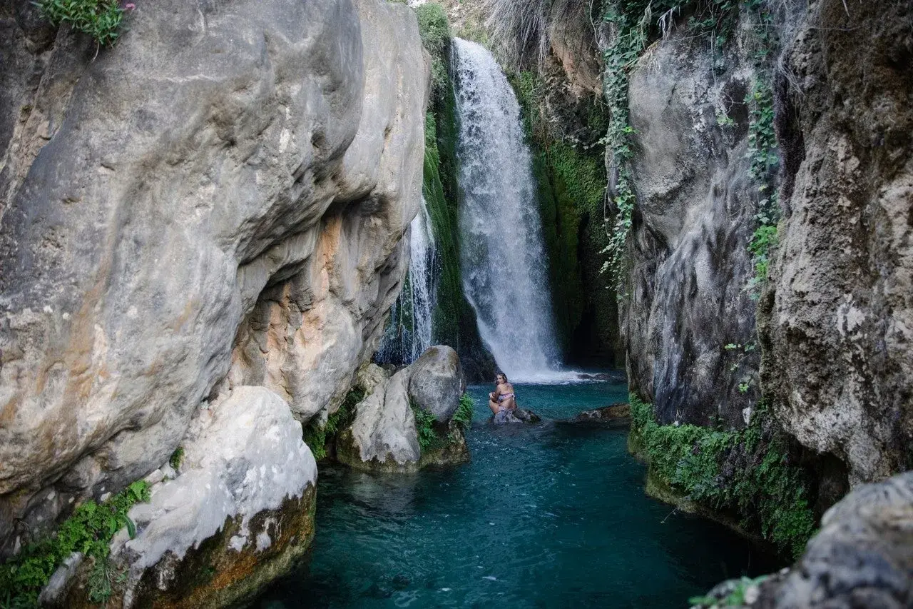 Una mujer se baña en las **fonts de Algar**, rodeada de rocas y vegetación, bajo una cascada de agua cristalina.