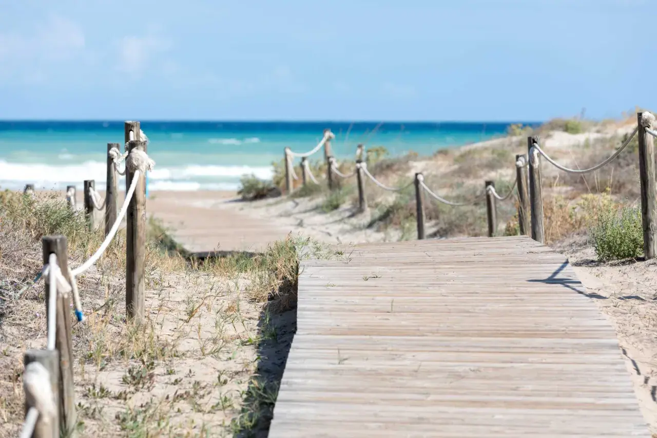 Pasarela de madera que conduce a la playa Raco de la Mar. El mar azul y las dunas de arena crean un paisaje sereno.