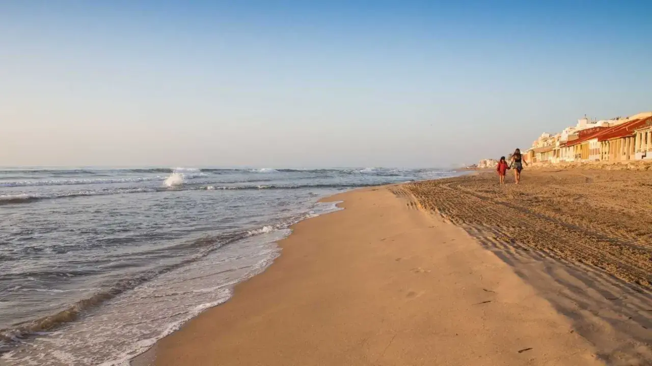 Dos personas pasean por la **playa Babilonia** al atardecer, con el mar y las casas de fondo.