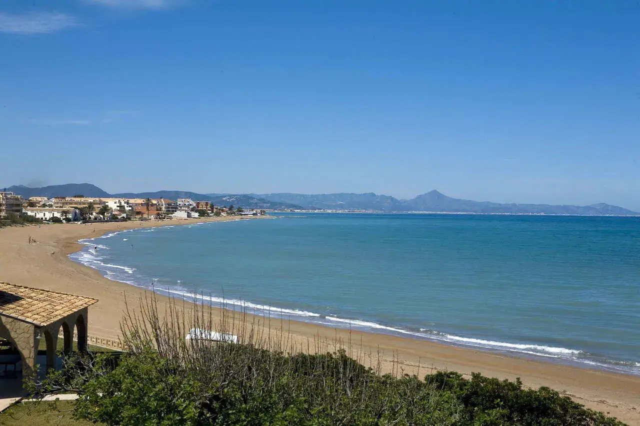 Amplia vista de la playa de Les Bovetes con arena dorada, mar azul y montañas al fondo. Un día soleado perfecto para disfrutar.