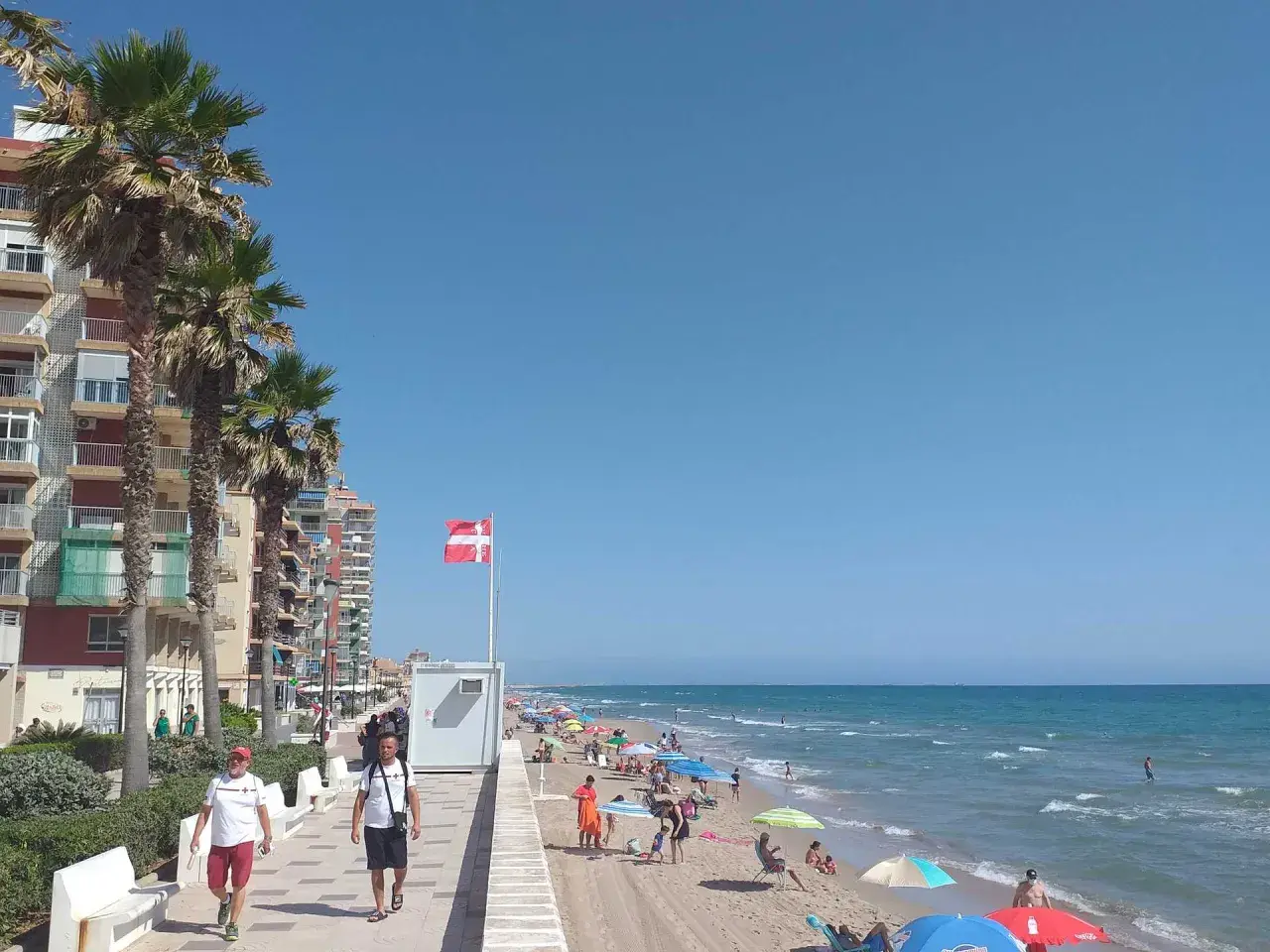 Gente disfrutando del sol y el mar en la playa Mareny de Barraquetes, con palmeras y edificios de apartamentos.