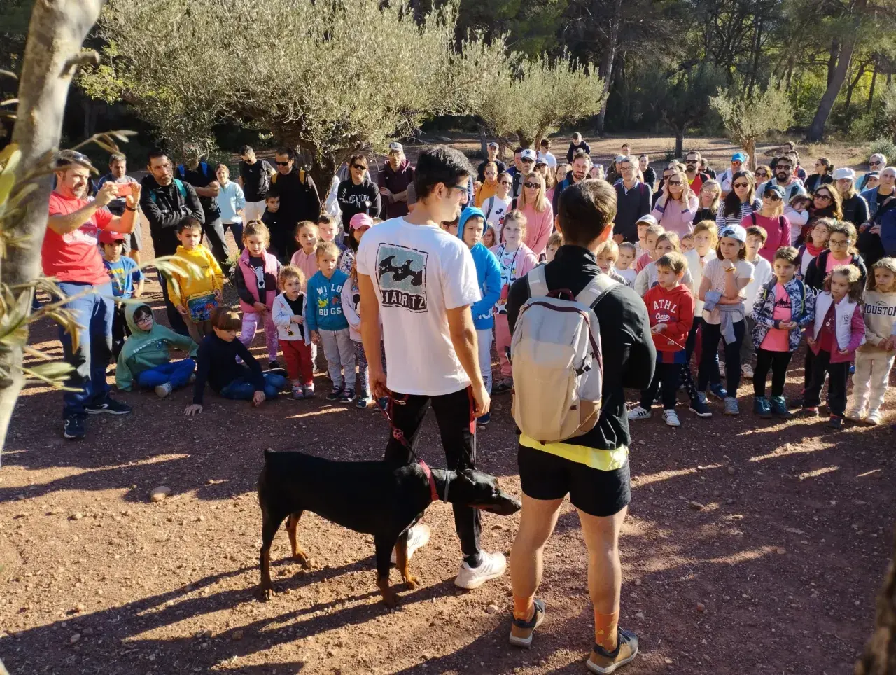 Un grupo de personas, incluyendo niños, se reúne en un sendero boscoso. Un hombre con una camiseta roja toma una foto con su móvil. Un perro Dóberman negro está atado a un joven. Quizás están buscando al **dragón de la Calderona**.