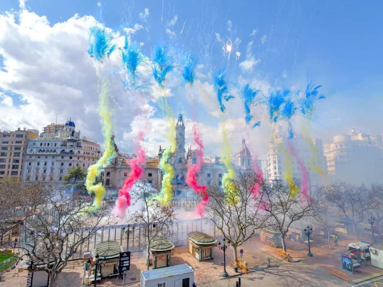 Explosión de color y ruido en la mascletà hoy. El cielo se tiñe de azul, amarillo y rosa sobre edificios históricos.
