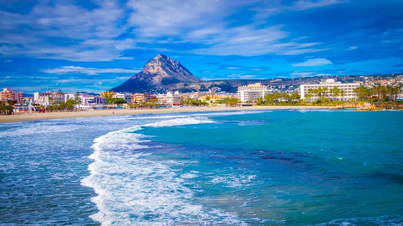 Olas turquesas rompen en la arena dorada de la playa del Arenal Javea, con edificios y el imponente Montgó al fondo.