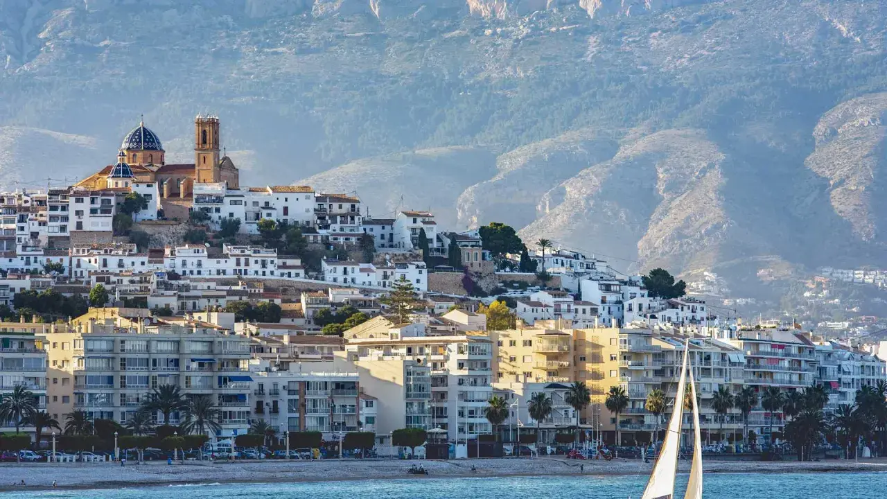 Vista del **castell de l'Olla** coronando la colina, con sus casas blancas y la iglesia de cúpula azul. Un velero navega frente a la costa.