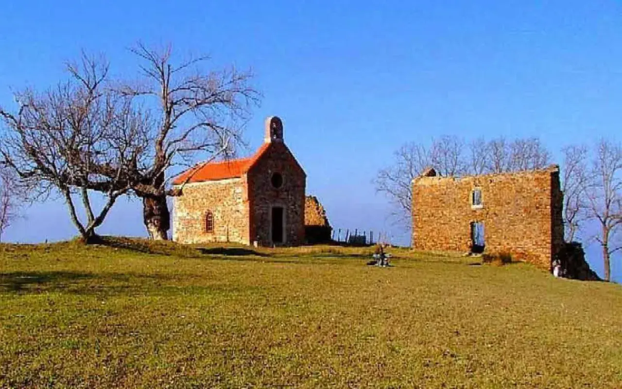 La ermita de Santa Catalina, una pequeña iglesia de piedra con tejado naranja, se alza junto a unas ruinas bajo un cielo azul. Un árbol desnudo y un paisaje verde completan la escena.