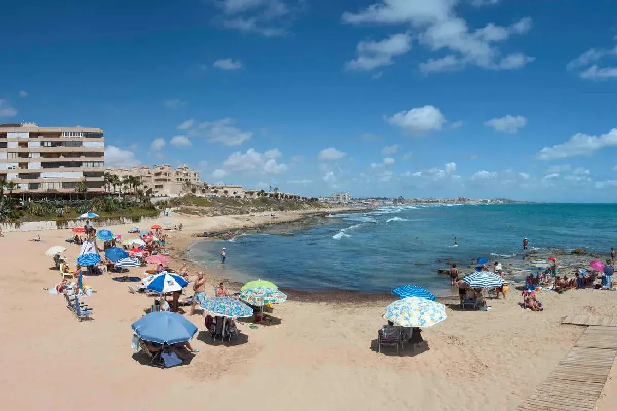 Gente disfrutando del sol y el mar en la playa Cabo Cervera, con sombrillas de colores y edificios al fondo.