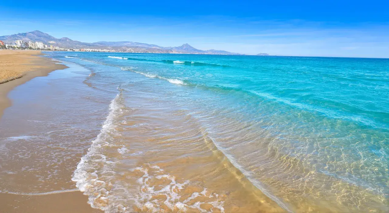 Aguas turquesas y arena dorada en las calas de San Juan, Alicante. El mar Mediterráneo acaricia la costa, con montañas al fondo.