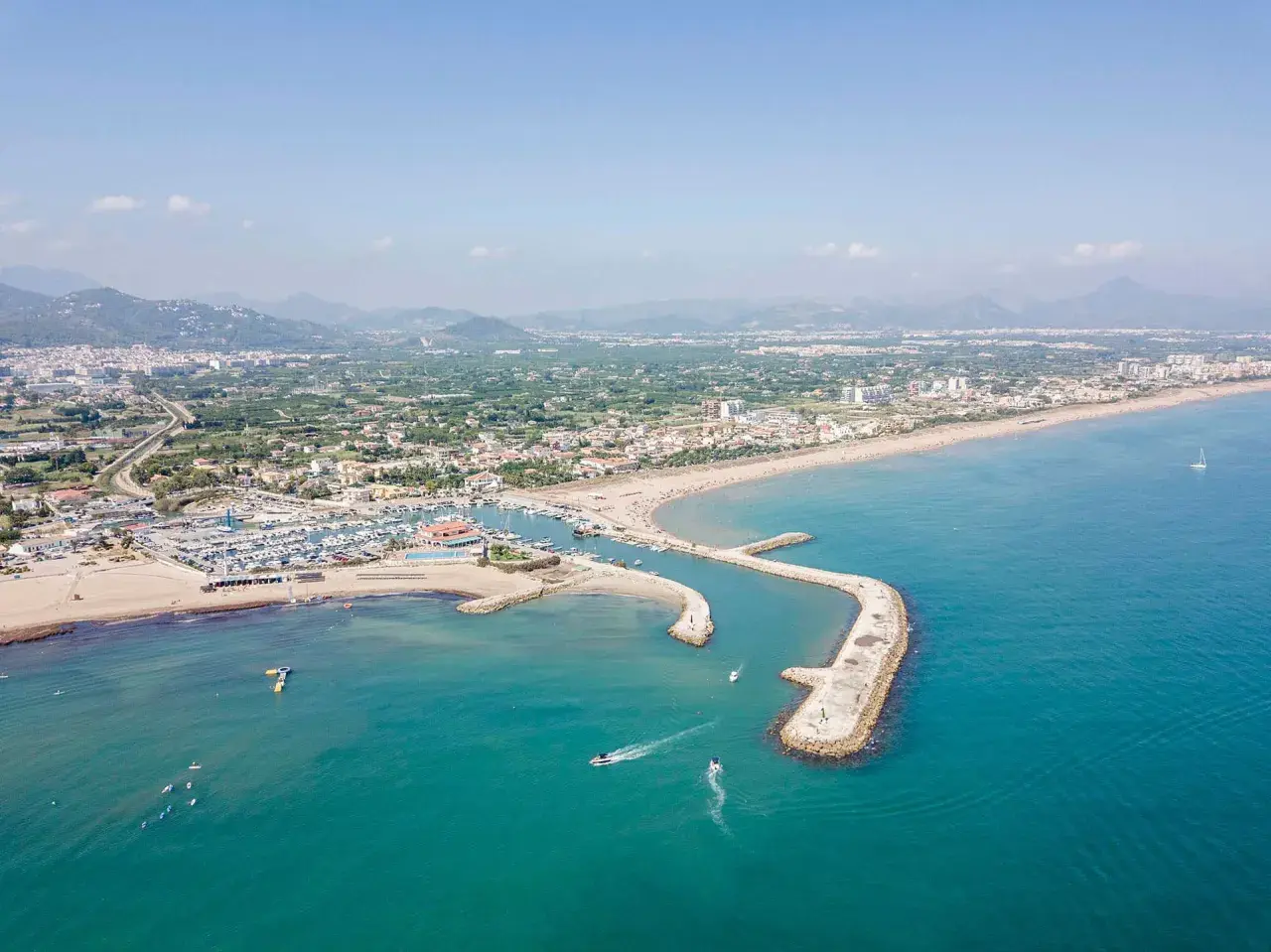 Vista aérea del puerto de Oliva con barcos navegando y la playa llena de gente.