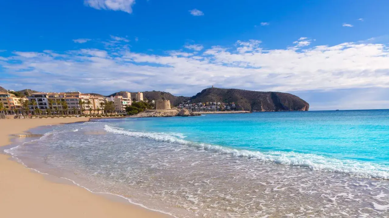 La playa de l'Ampolla en Moraira, con su arena dorada y aguas turquesas, invita a un día de sol y mar. Al fondo, edificios y una torre defensiva se integran en el paisaje.