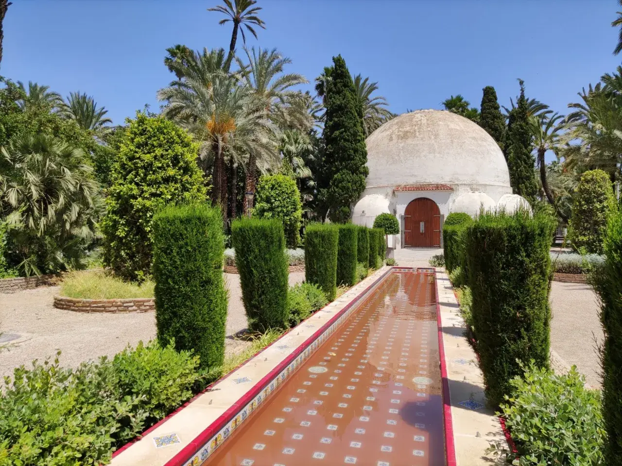 Un estanque de agua rojiza y un edificio abovedado en el parque el palmeral, rodeado de palmeras y vegetación exuberante.