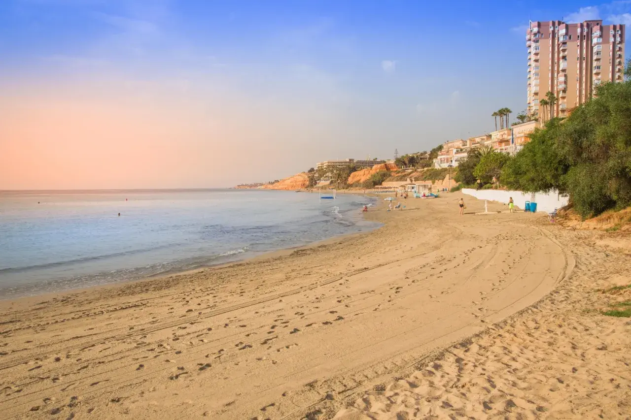Playa con arena dorada y el mar azul. Al fondo, un barranco rubio con edificios y vegetación.