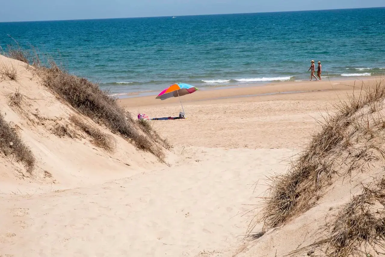 Dunas de arena y mar azul en el mareny de Sant Llorenç. Una sombrilla colorida y dos personas pasean por la orilla.