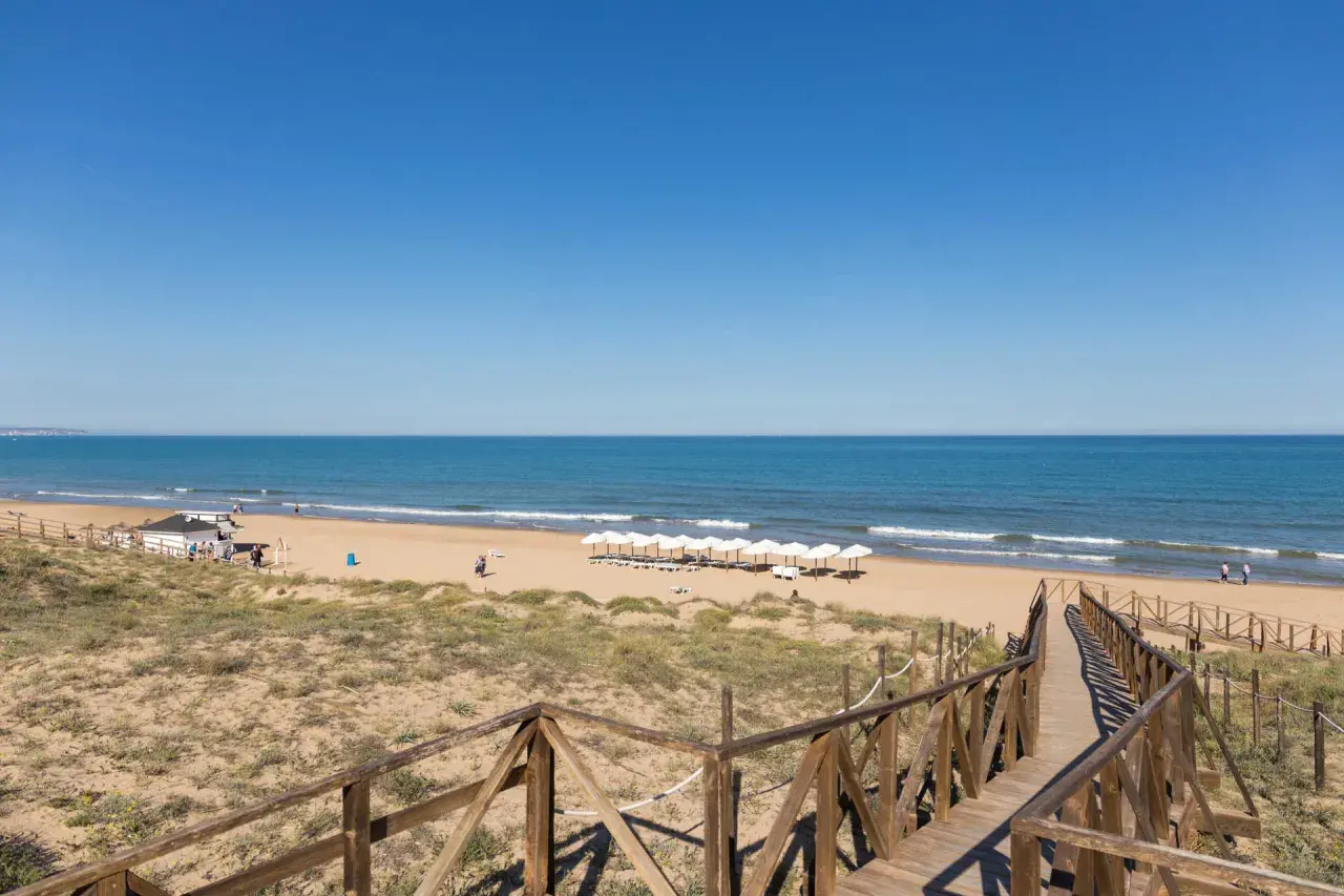 Pasarela de madera hacia la playa Moncayo, con arena dorada, mar azul y sombrillas blancas alineadas.